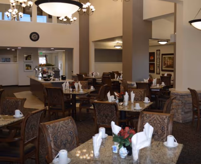 Interior view of a dining room in a senior living facility with multiple tables set with white napkins, cups, and small flower arrangements. The room features patterned chairs, chandeliers, and warm lighting with beige walls and stone accents on the columns.