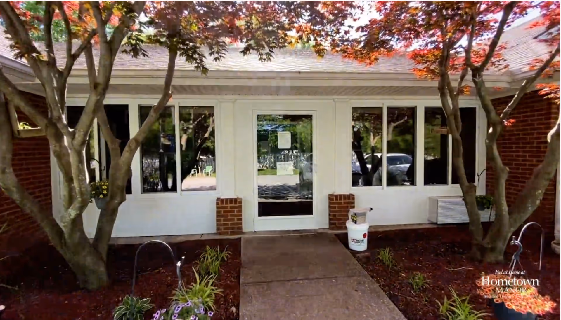 Entrance to a building with a glass door and windows on either side, surrounded by red brick walls and trees with red leaves. There are plants and flowers in the garden beds along the walkway leading to the door.