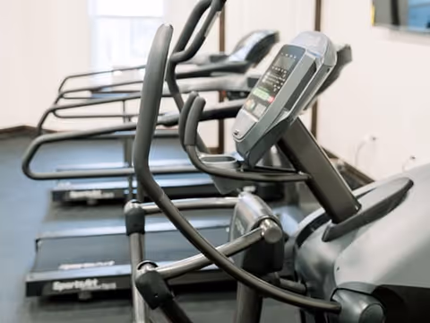 Row of elliptical machines and treadmills in a fitness room with large windows allowing natural light.
