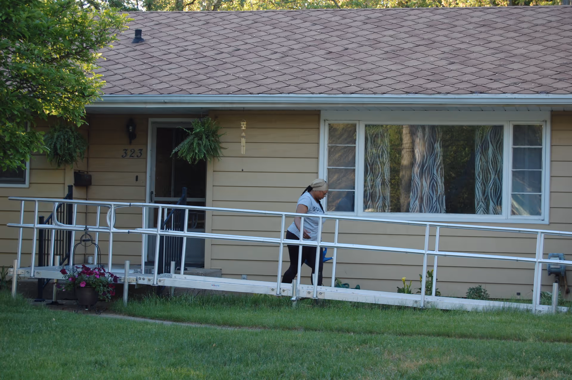 A woman walking on a metal wheelchair ramp leading to the entrance of a single-story beige house with a brown shingled roof. The house has a large window with patterned curtains and hanging plants near the door. The lawn in front is green and well-kept.