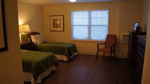 A simple bedroom with two twin beds with green bedspreads, a nightstand and lamp, a window with blinds, a chair, and a dresser.