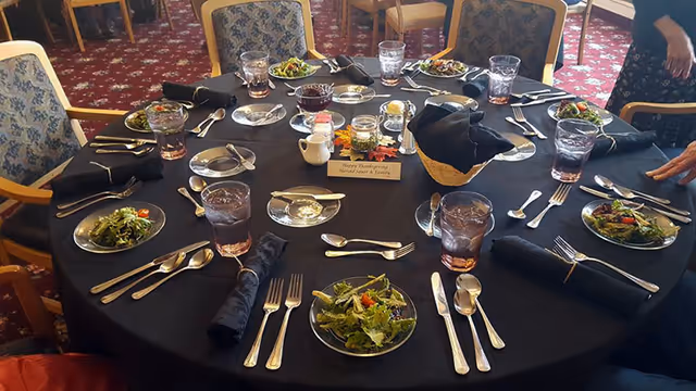 A round dining table set for eight people with black tablecloth, each place setting having a salad plate with greens, a glass of water, silverware, and a black cloth napkin. In the center of the table are condiments and a small sign that reads 'Happy Thanksgiving'. Chairs with patterned upholstery surround the table, and part of a person is visible on the right side of the image.