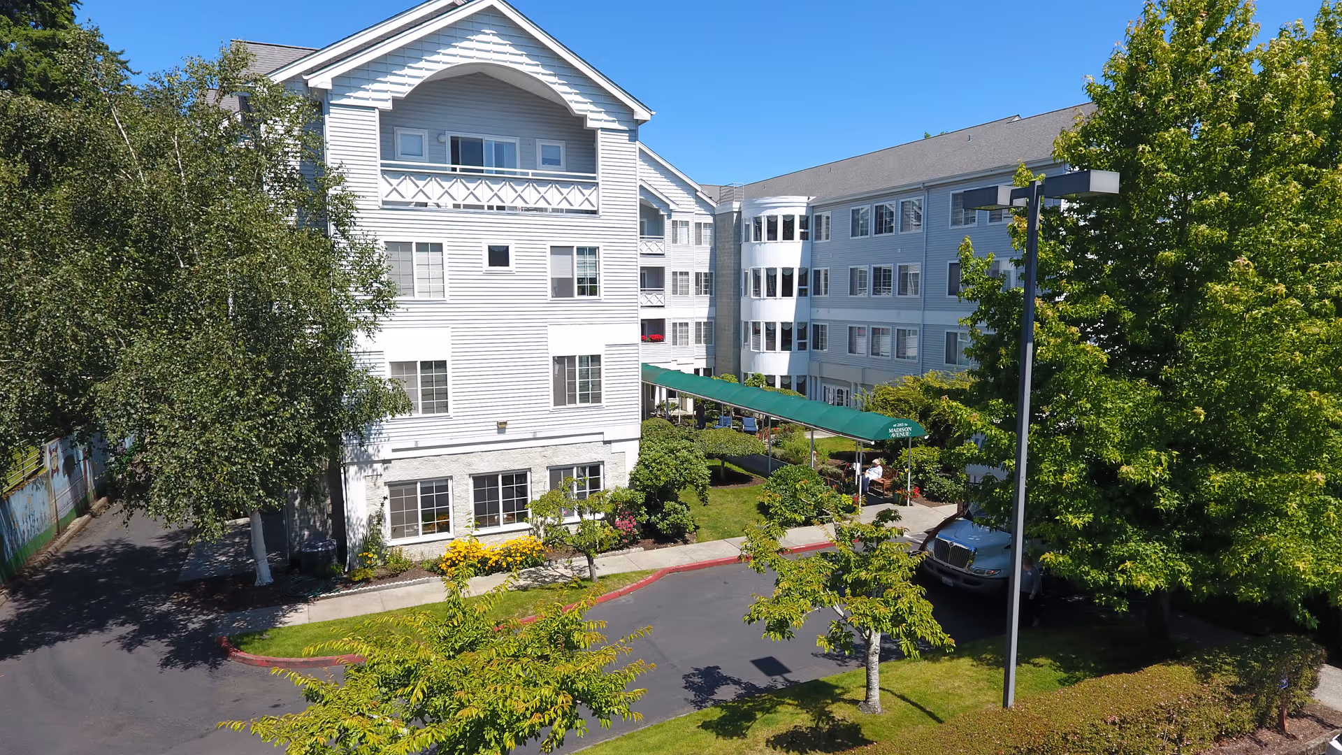 Exterior view of a multi-story white senior living facility building with multiple windows, surrounded by green trees and landscaping under a clear blue sky. A covered entrance with a green canopy is visible, along with a driveway and parked vehicle.