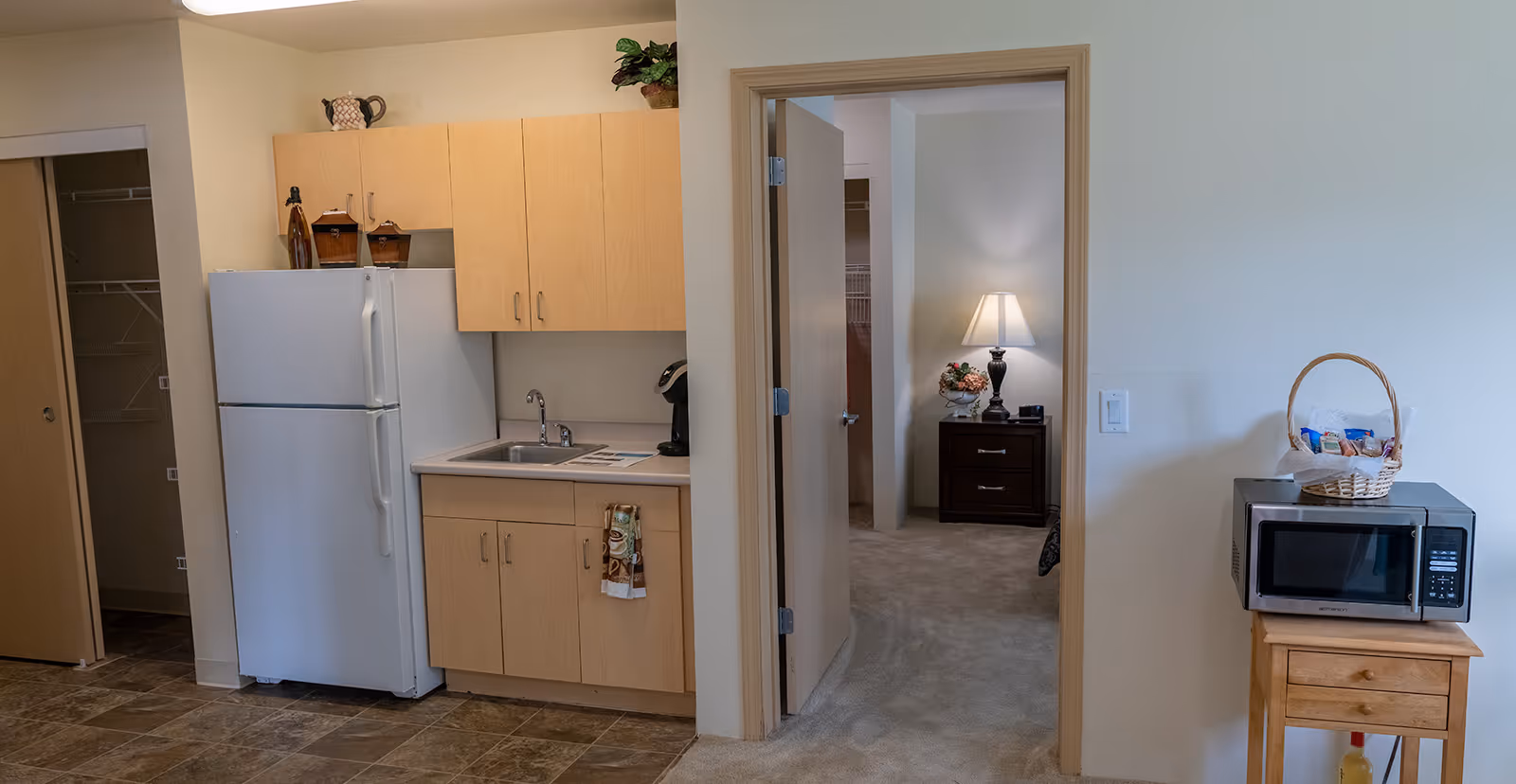 Interior view of a small kitchen area with light wood cabinets, a white refrigerator, a sink, and a coffee maker. To the right, there is a microwave on a small wooden table with a basket of snacks on top. An open doorway leads to a carpeted bedroom with a nightstand, a lamp, and a floral arrangement.