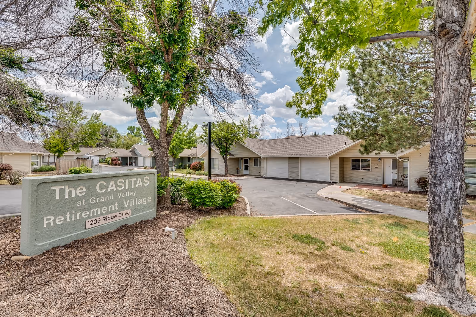 Exterior view of The Casitas at Grand Valley Retirement Village showing a sign with the address 1209 Ridge Drive, surrounded by trees and shrubs, with single-story residential buildings in the background under a partly cloudy sky.