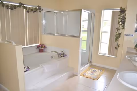 Bright bathroom featuring a large white bathtub with a towel draped over the edge, a glass-enclosed shower, two sinks with mirrors, beige tiled floor, and vertical blinds on the windows. Decorative plants and flowers are placed around the room.
