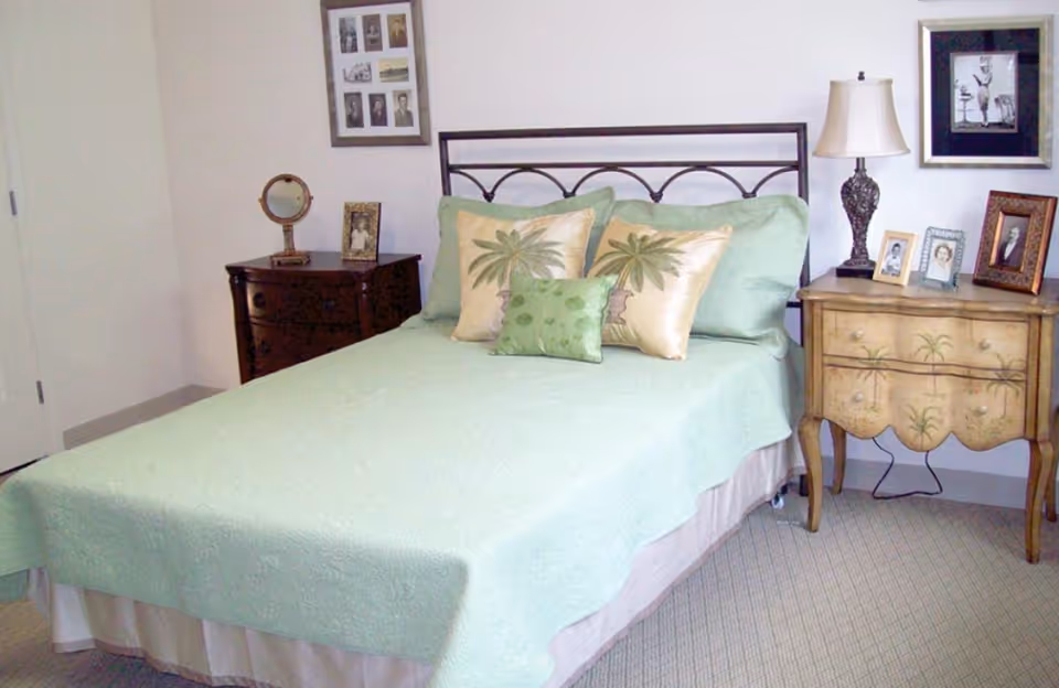 A bedroom with a bed covered in a light green bedspread and three decorative pillows with palm tree designs. There are two nightstands on either side of the bed, one dark wood with a round mirror and picture frame, and the other a lighter wood with a lamp and several framed photos. The walls are light-colored with framed pictures hanging above the nightstands.