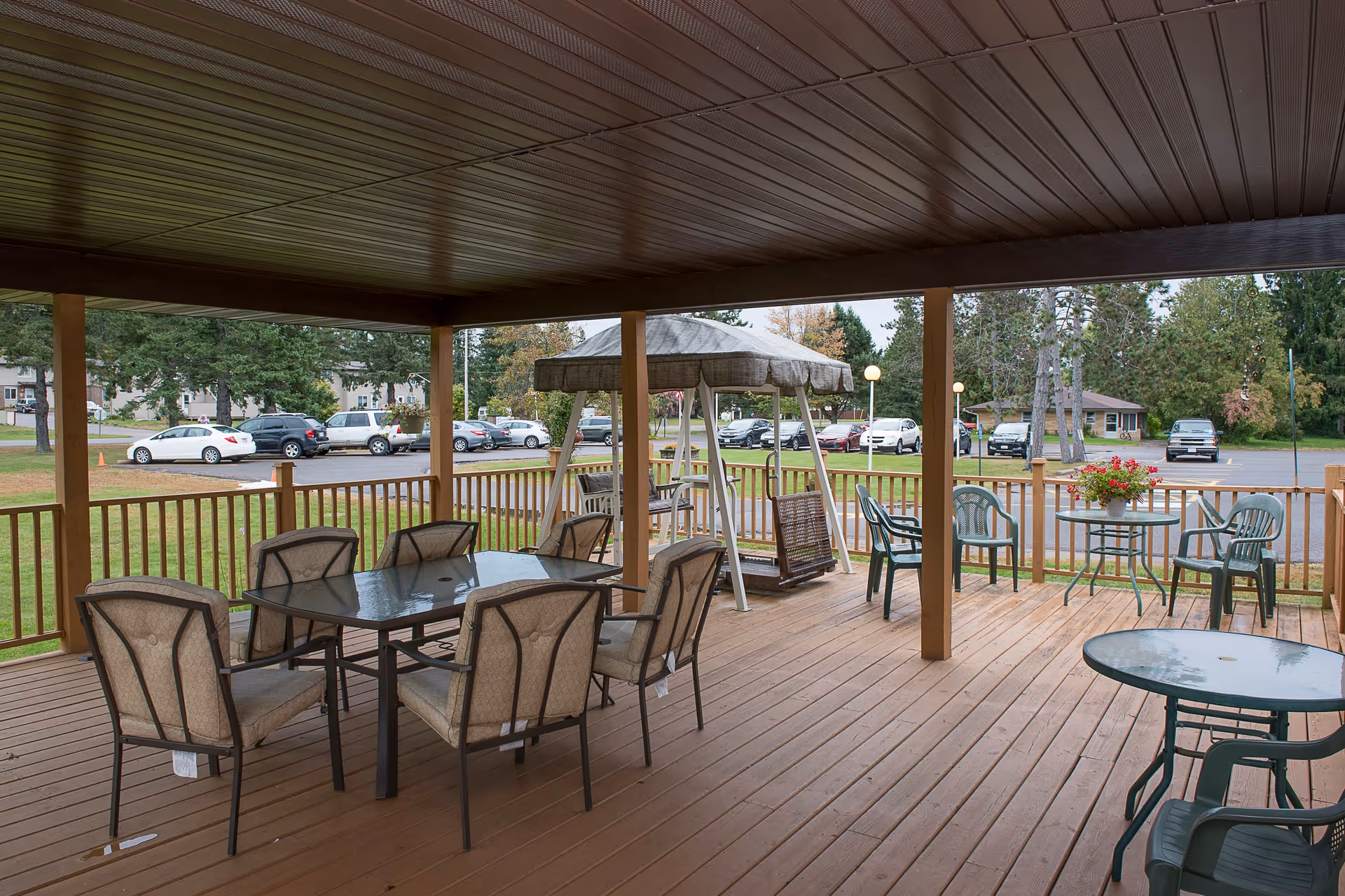 Covered outdoor patio area with multiple seating arrangements including a glass-top table with six cushioned chairs, a swing with a canopy, and two smaller round tables with plastic chairs. The patio overlooks a grassy area and a parking lot with several cars parked. Trees and buildings are visible in the background.