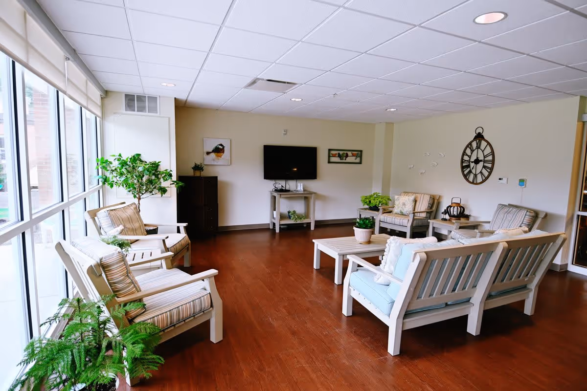 Bright communal seating area with couches, chairs, plants and a wall-mounted TV in a senior living facility.