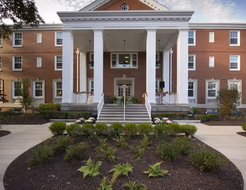 Front facade of a red-brick senior living building featuring a portico with white columns, steps, and landscaped beds.