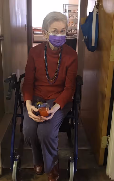 An elderly woman wearing a purple face mask, red sweater, and beaded necklaces is sitting on a walker in a narrow hallway, holding a glass of red liquid. The background shows a room with shelves and various items.