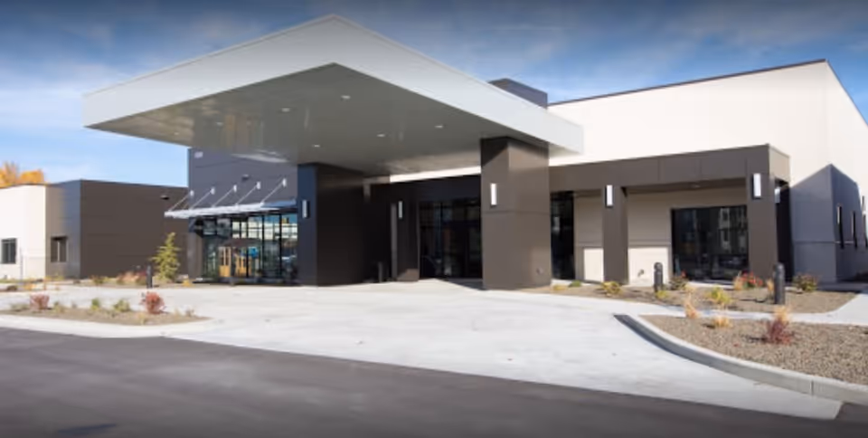 Exterior view of a modern senior living facility building with a large covered entrance, glass doors, and landscaped areas with small plants and shrubs.