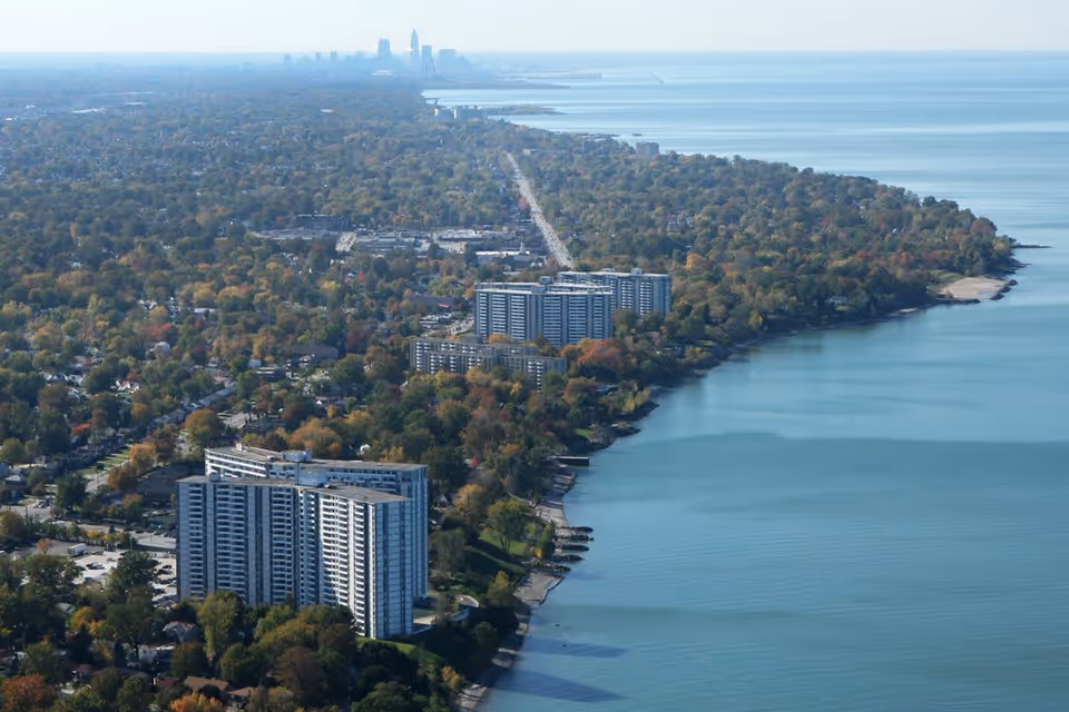 Aerial view of a lakeside urban area with several high-rise buildings near the shoreline, dense tree coverage, and a city skyline visible in the distance under a clear sky.