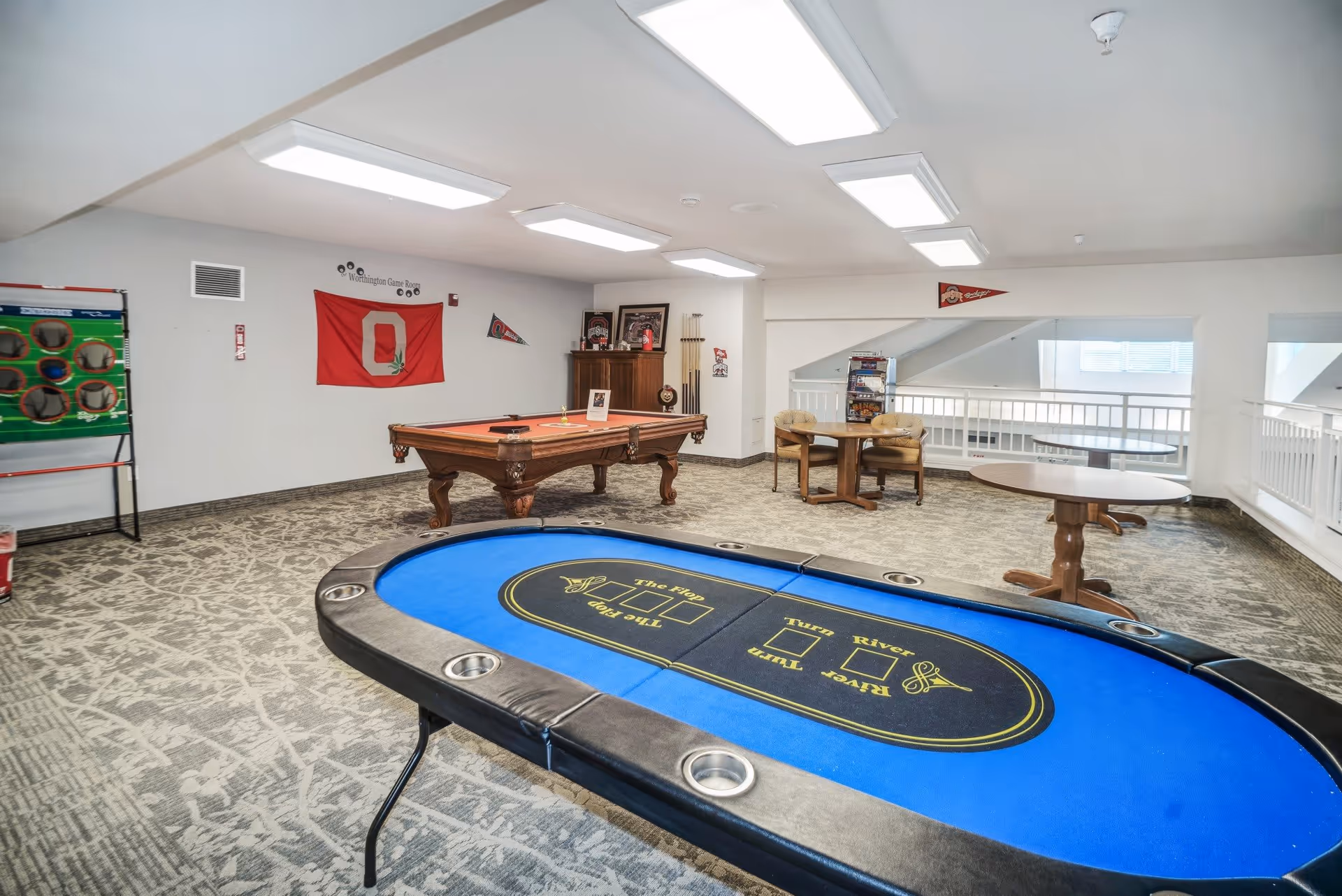 A recreational room with a poker table in the foreground, a pool table in the middle, and a small seating area with two chairs and a round table near the back. The walls are decorated with sports pennants and a red flag with a large letter O. The room has bright overhead lighting and carpeted flooring.