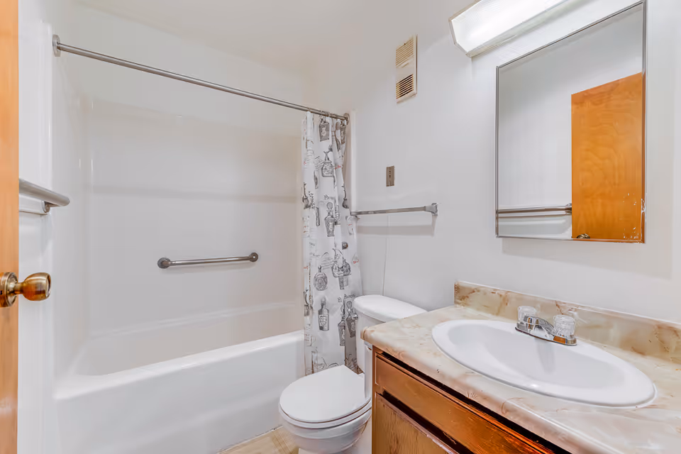 A small bathroom with a white bathtub and shower combination, a white toilet, and a sink with a marble-patterned countertop. There is a mirror above the sink and a shower curtain with a decorative pattern. The walls are white, and there is a wooden door partially visible.