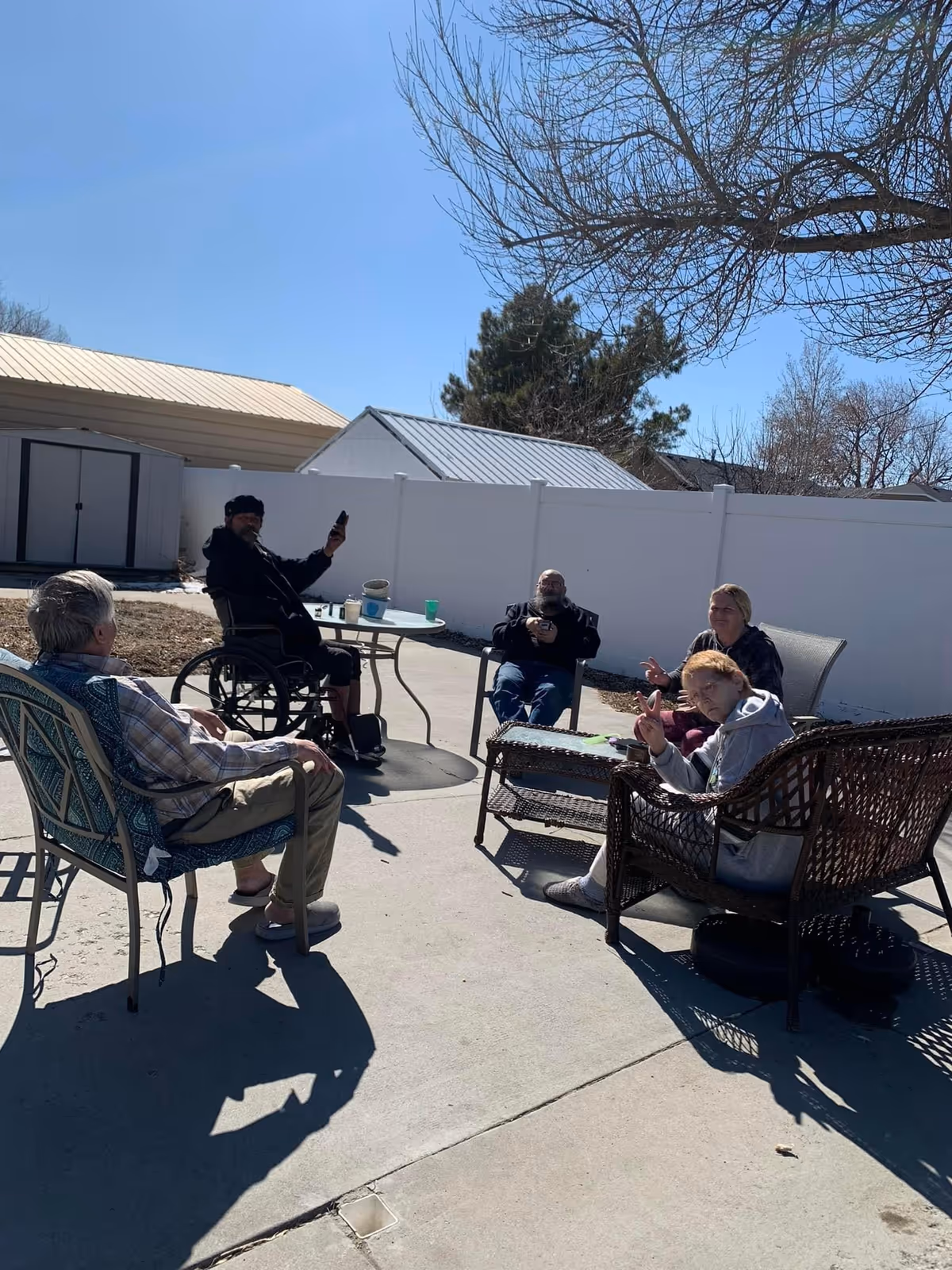 Five people sitting outdoors on patio furniture and chairs in a fenced backyard area on a sunny day. One person is in a wheelchair holding up a phone, and another person is making a peace sign towards the camera. There are trees and small sheds visible in the background.
