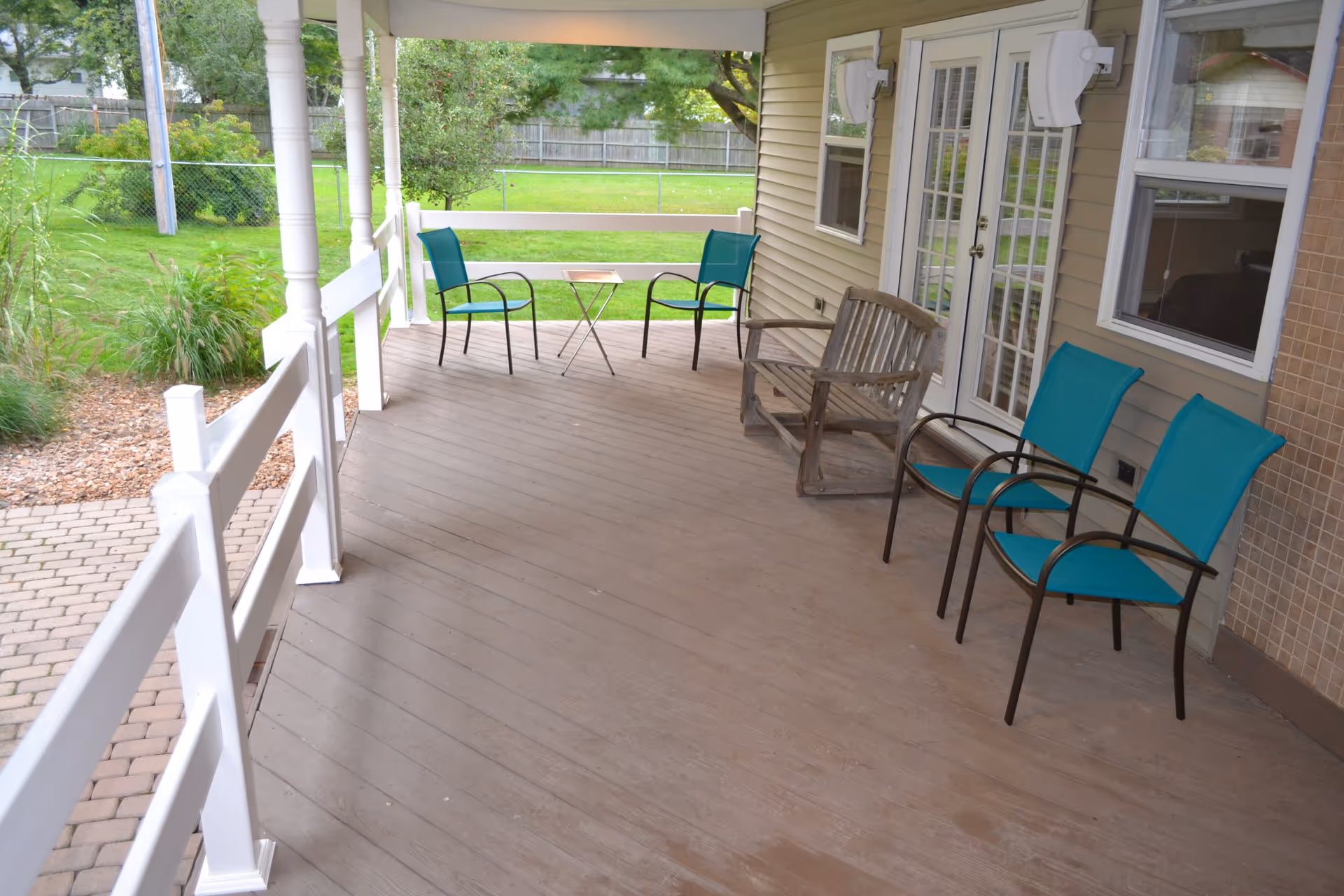 Covered outdoor porch area with wooden flooring, white railing, and several chairs including two turquoise chairs, a wooden bench, and a small table. The porch overlooks a green lawn with trees and a fence in the background.