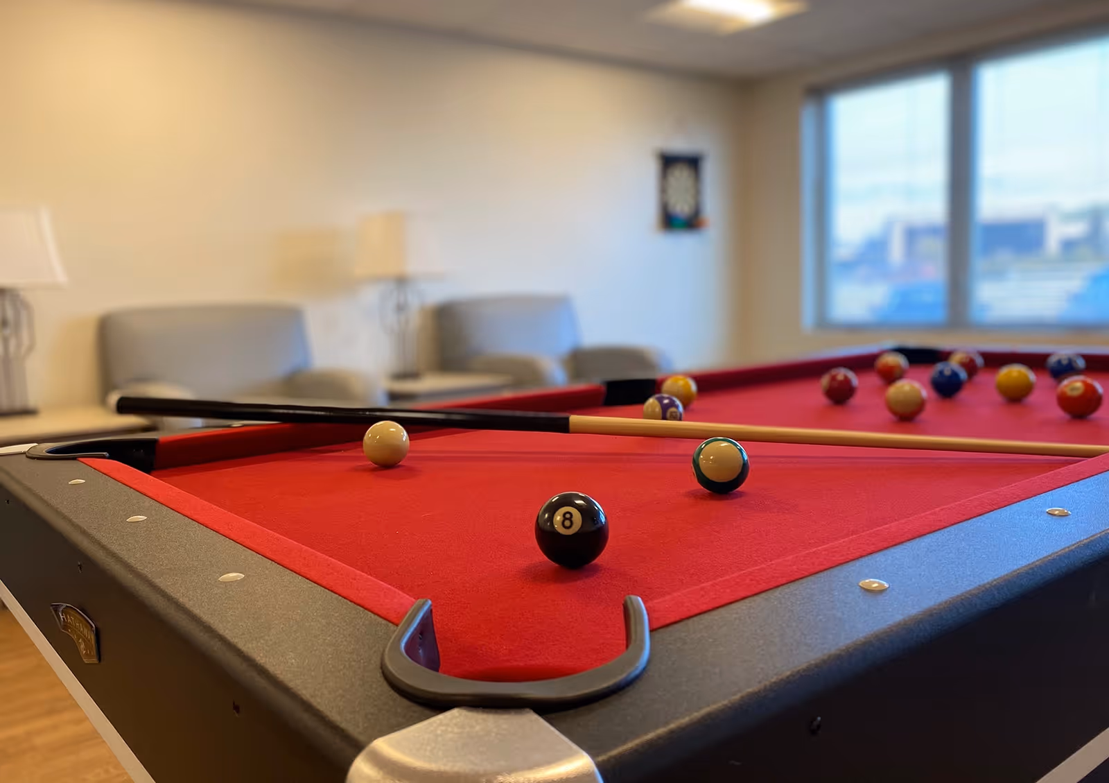 A red felt pool table with billiard balls and a cue stick resting on it, set in a room with beige walls, two gray armchairs, a floor lamp, and a window showing an outside view.