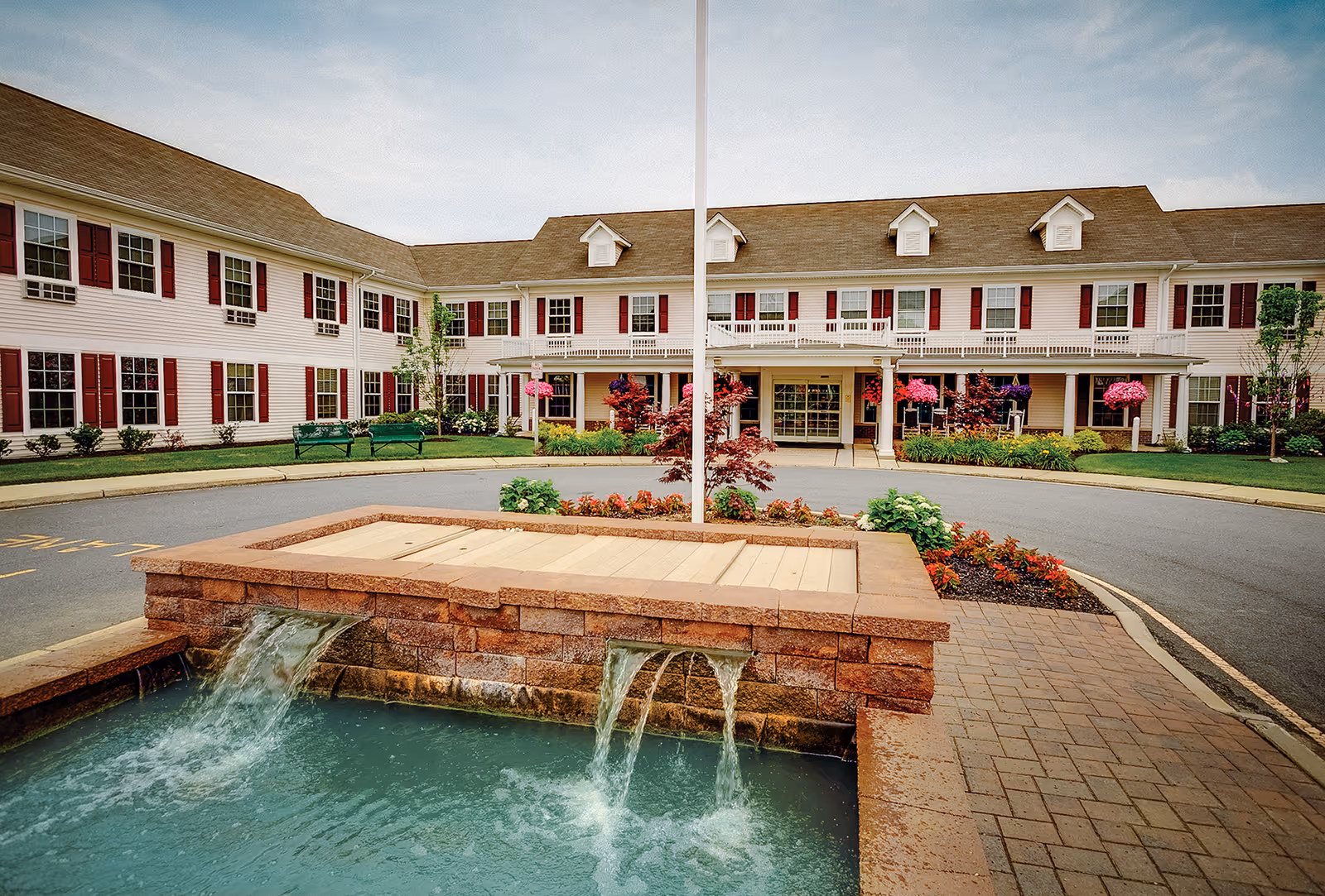 Front exterior view of Brandywine Toms River by Monarch, showing a two-story building with white siding and red shutters. In front of the building is a circular driveway with a landscaped area featuring a water fountain with flowing water, colorful flowers, and greenery under a partly cloudy sky.