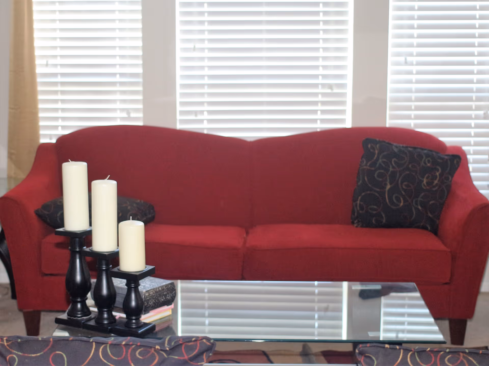 A red upholstered sofa with two decorative black pillows featuring a swirl pattern, positioned in front of a window with white blinds. In front of the sofa is a glass coffee table with three white pillar candles on black candle holders and a stack of books.