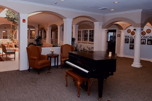 Interior view of a senior living facility common area featuring a black grand piano with a matching bench, two brown armchairs with a small round table between them, white columns and arches, and a wall with framed photos and plaques in the background.