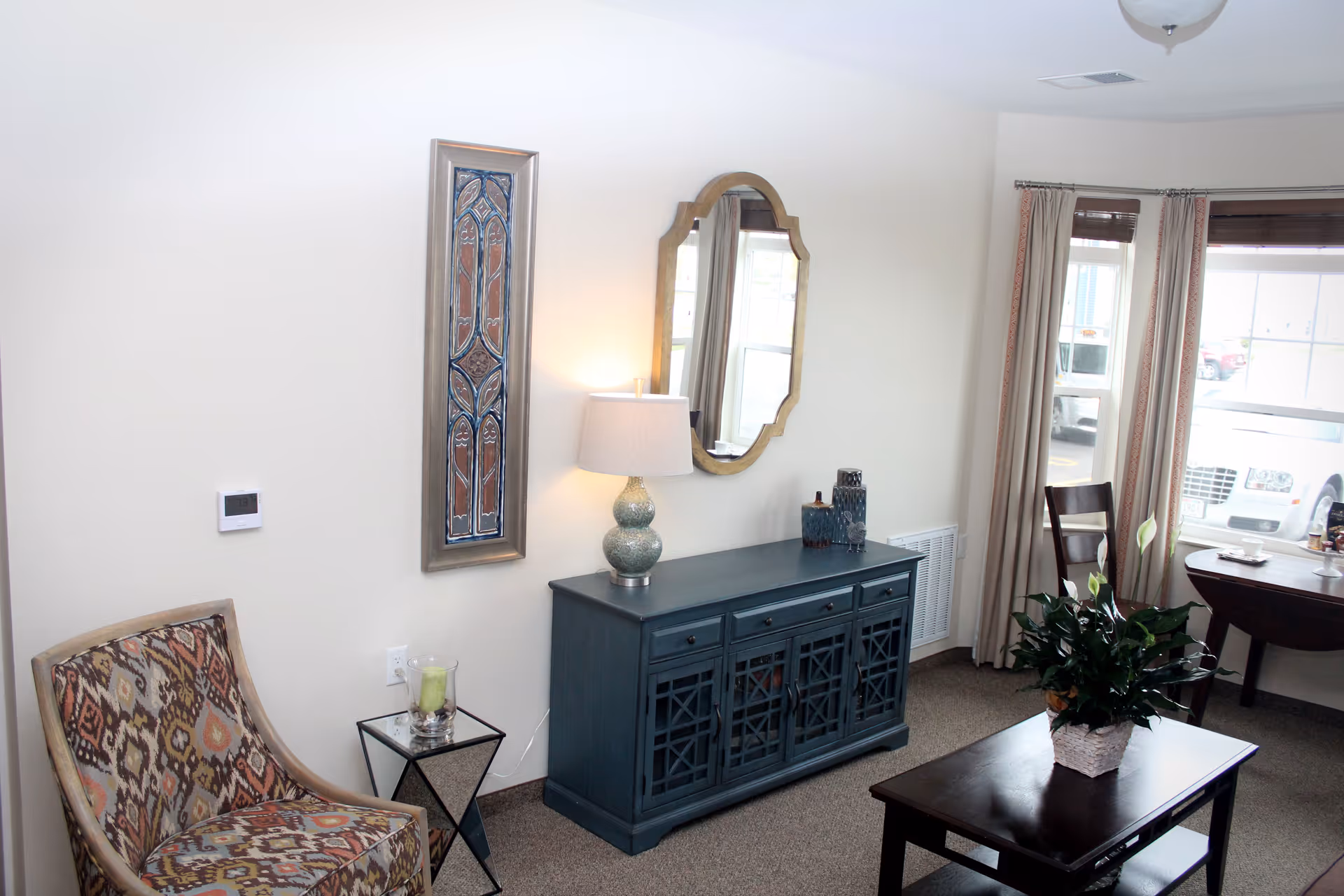 Well-lit living room with a patterned armchair, a blue console table with a lamp and mirror, a coffee table with a potted plant, and windows with curtains.
