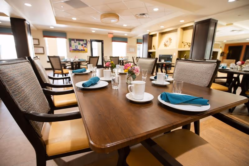 A dining room with a wooden table set for a meal, featuring white plates, cups, glasses, and teal cloth napkins. The room has multiple tables and chairs, soft lighting, and decorative flowers on the tables. The background shows windows, artwork, and a cozy interior setting.