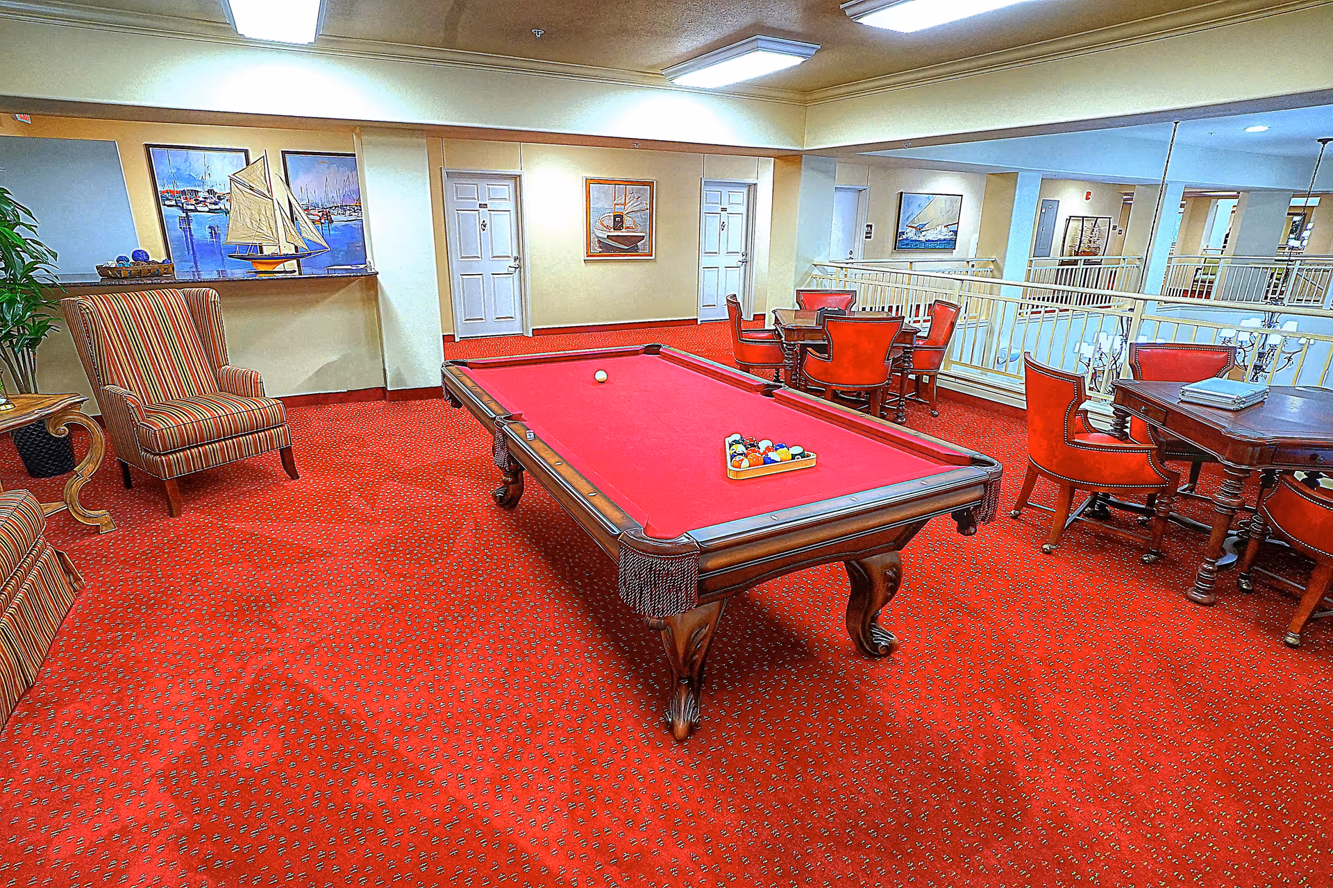Indoor lounge with a red-felt pool table on red carpet surrounded by chairs and tables in a senior living facility.