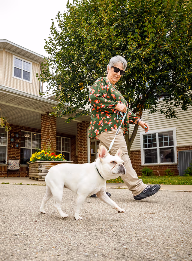 An elderly woman wearing sunglasses and a green floral blouse walks a white French bulldog on a leash outside a senior living facility with brick and siding exterior and a tree in the background.
