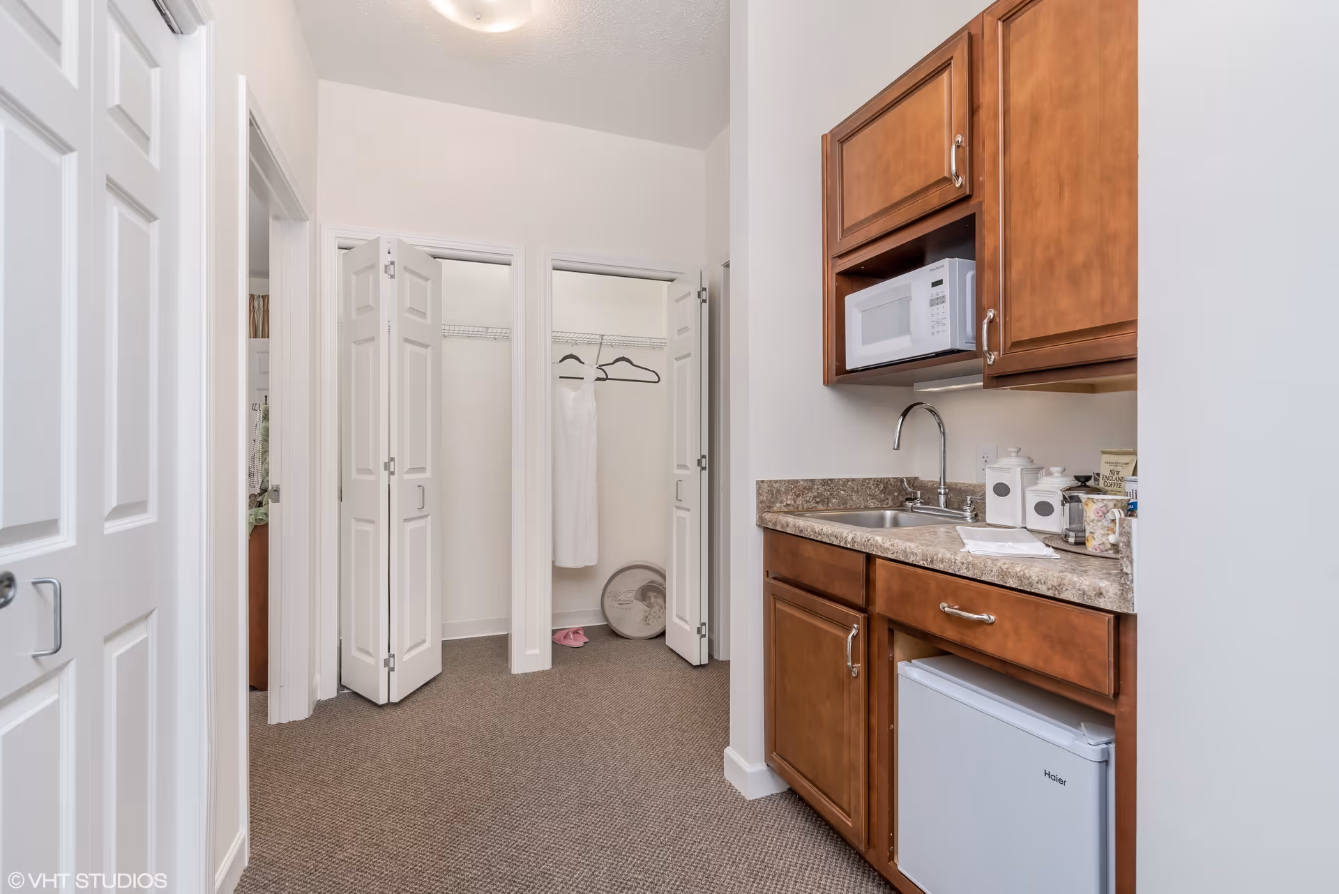 Interior view of a small kitchenette area with wooden cabinets, a countertop with a sink, a microwave, and a mini refrigerator. To the left, there are two open closets with white bi-fold doors, one containing a white dress hanging on a hanger and a pair of pink slippers on the floor. The walls are white and the floor is carpeted in a neutral color.