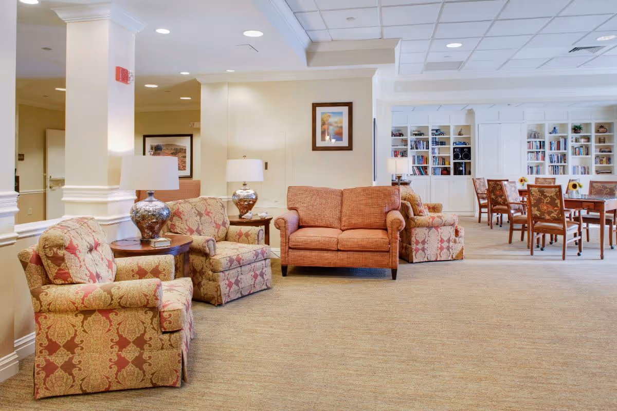 A spacious and well-lit senior living common area featuring patterned armchairs and a sofa arranged around wooden side tables with decorative lamps. In the background, there are bookshelves filled with books and a dining area with tables and chairs, creating a cozy and inviting atmosphere.