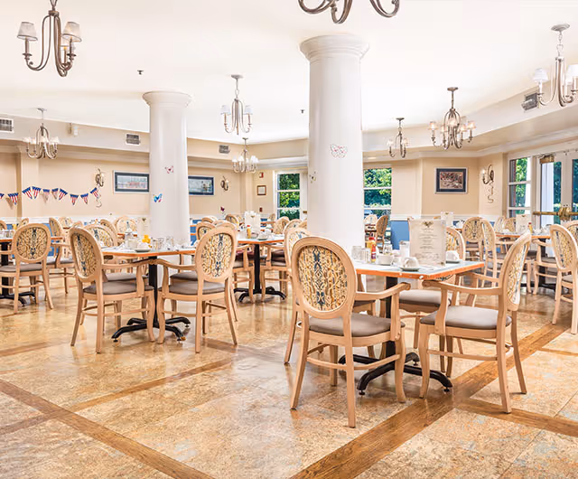 A spacious dining room with multiple wooden tables and chairs arranged neatly. The chairs have cushioned seats and decorative backs. The room features large white columns, chandeliers hanging from the ceiling, and windows letting in natural light. The tables are set with cups, plates, and menus, ready for guests.