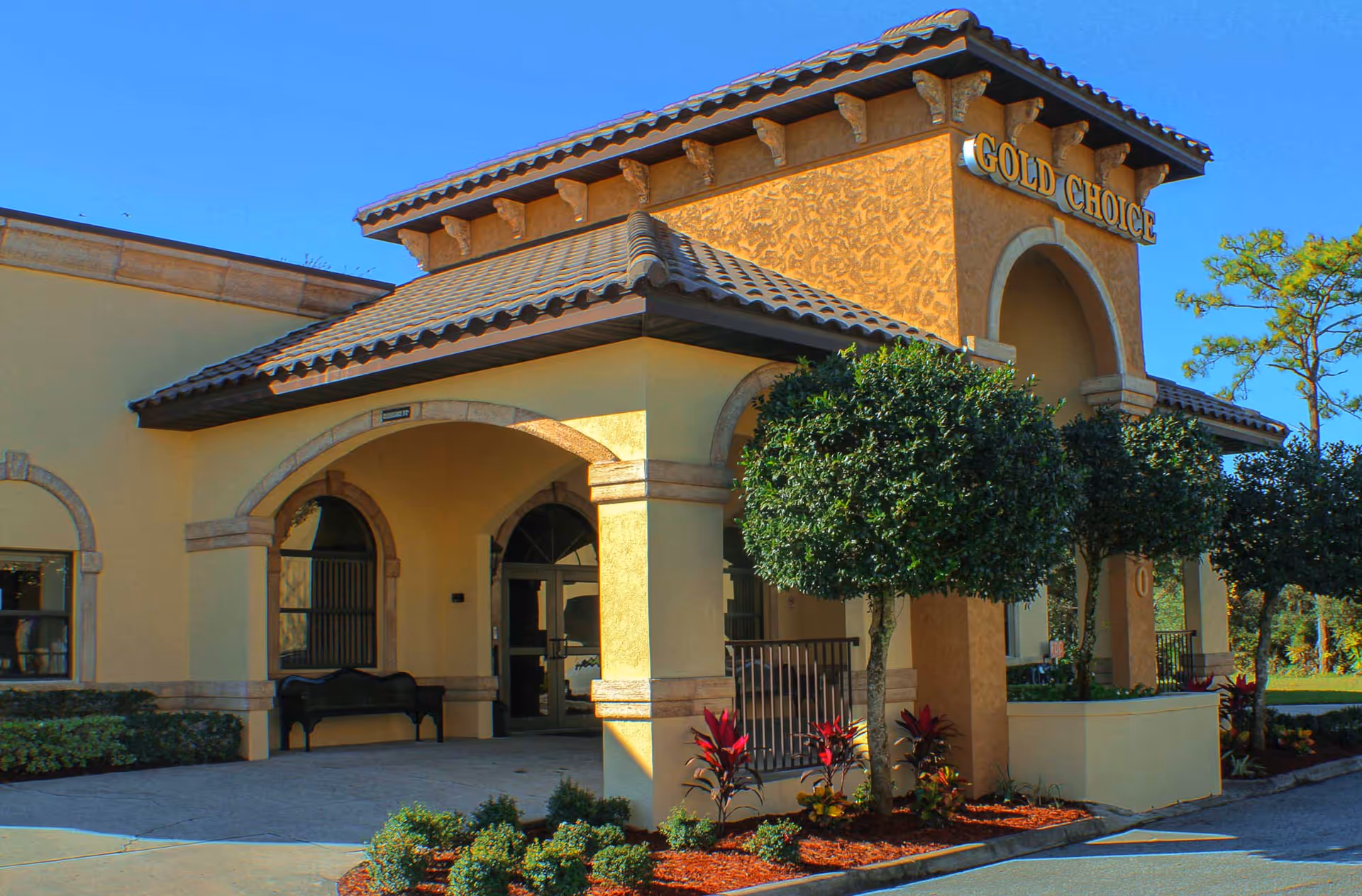 Exterior view of the Gold Choice Ormond Beach Assisted Living facility showing the building entrance with arched doorways, a tiled roof, manicured bushes, and a clear blue sky.