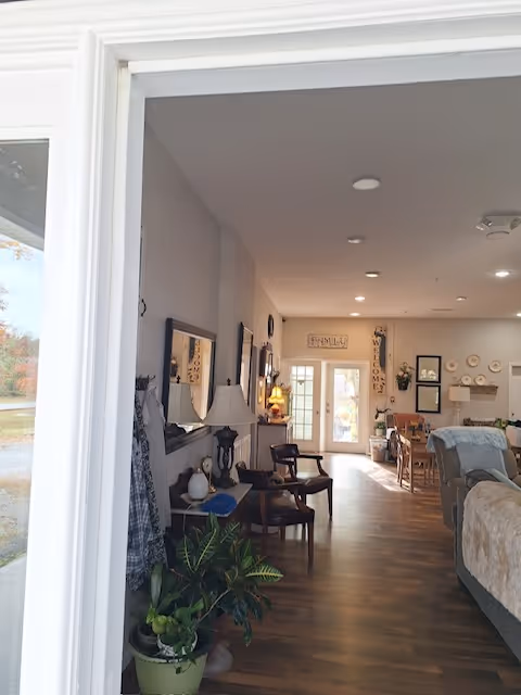 View through a doorway into a cozy living area with wooden flooring, two wooden chairs, a table lamp, potted plants, and wall decorations including framed pictures and a 'WELCOME' sign near a glass door letting in natural light.