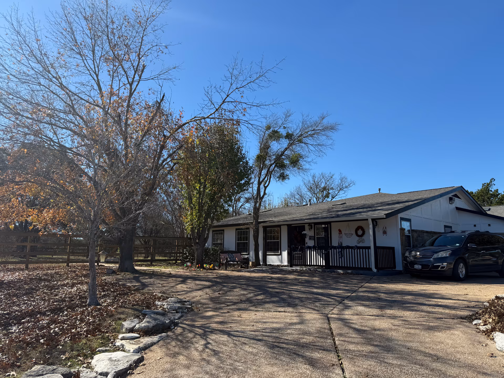 Exterior view of a single-story building with a driveway and parked car. The building has a small porch with a railing and some decorations on the wall. There are leafless trees and a wooden fence surrounding the property under a clear blue sky.