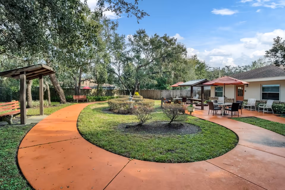 Outdoor courtyard area with a curved orange walkway, benches, small bushes, and tables with umbrellas. There are trees and a building with windows and chairs in the background under a partly cloudy sky.