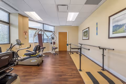 Small interior fitness and therapy room with exercise machines, a weight station, and parallel walking bars near windows.