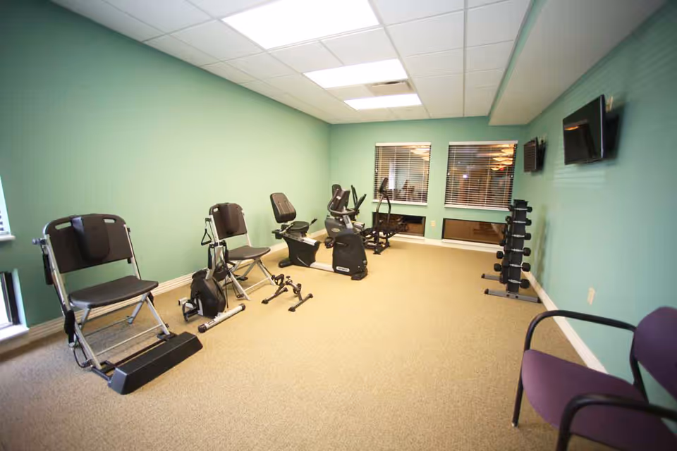 A small exercise room with light green walls and beige carpet. The room contains several pieces of exercise equipment including two recumbent exercise bikes, a pedal exerciser, and a set of dumbbells on a rack. There are two windows with blinds and two wall-mounted televisions. A purple chair is visible in the foreground.