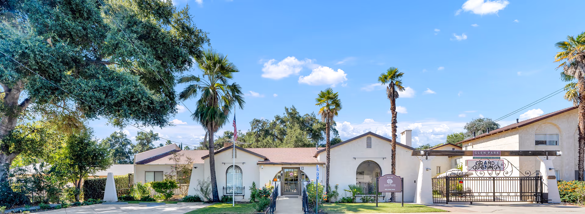 Exterior view of Glen Park at Ojai senior living facility on a sunny day with a clear blue sky. The building is white with a brown roof and has a gated entrance with a sign that reads 'Grand Opening'. There are palm trees and other greenery around the building.