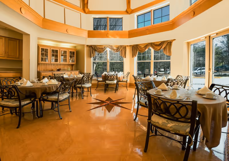Bright, elegant dining room with round tables set for meals, wrought-iron chairs, large windows, and a high curved ceiling.