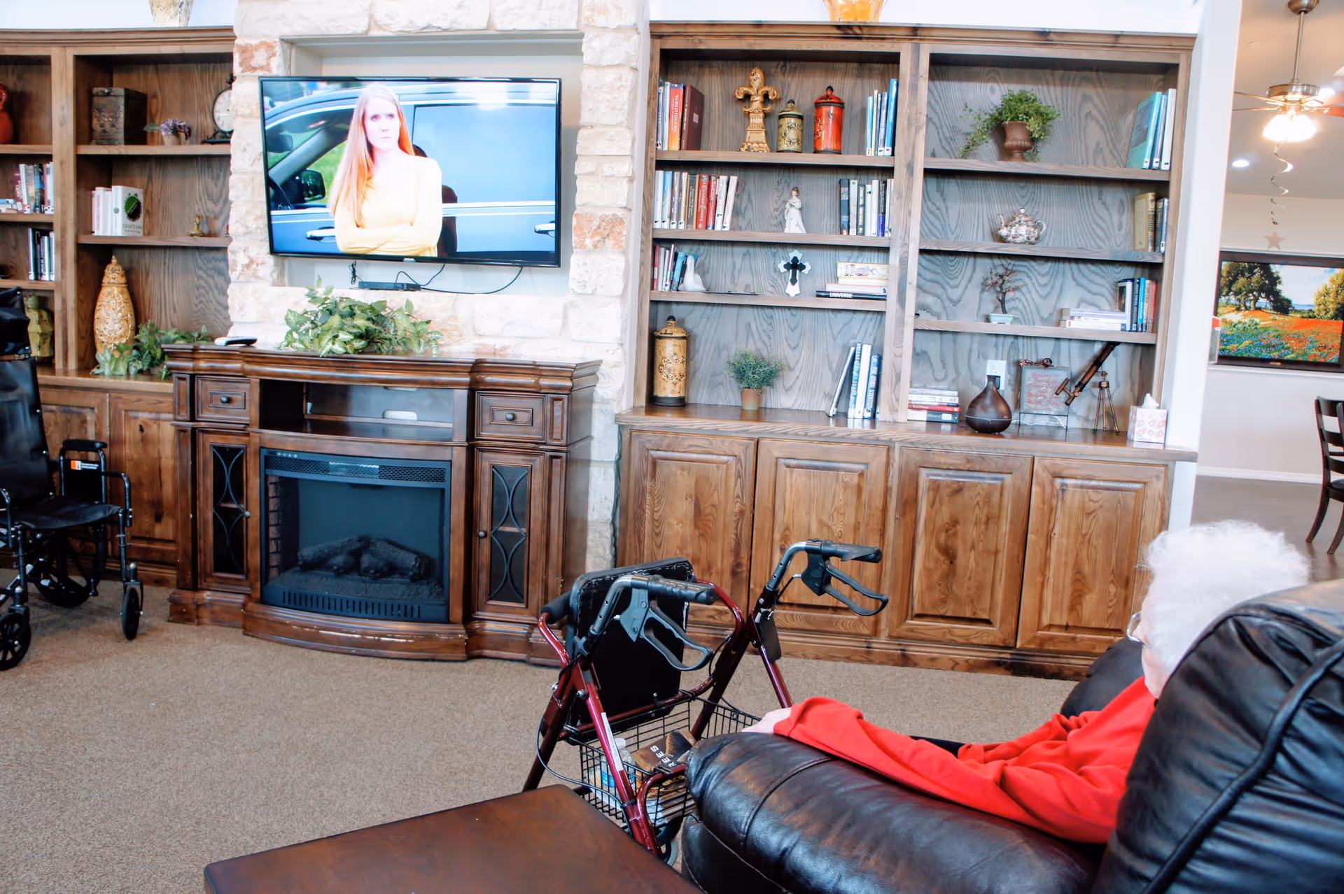 An elderly person with white hair wearing a red sweater sits on a black leather chair watching a television mounted on a stone wall above a wooden electric fireplace. The room features wooden built-in bookshelves filled with books and decorative items. A walker is positioned next to the chair, and another wheelchair is visible to the left. The space appears cozy and well-lit with a ceiling fan and a dining area visible in the background.