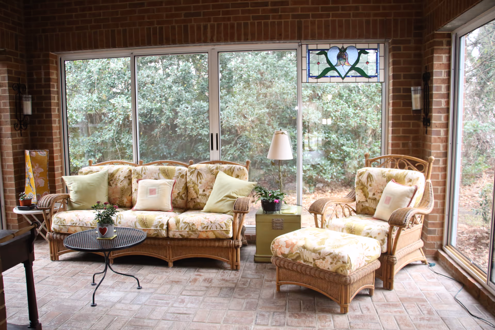 A cozy sunroom with wicker furniture including a floral-patterned sofa, armchair, and ottoman. The room has large sliding glass doors and windows showing greenery outside. There is a small round metal table with a potted plant, a green side table with a lamp and another plant, and brick walls and floor.