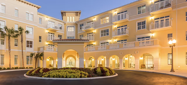 Exterior view of a large, multi-story senior living facility building at dusk with warm lighting. The building features balconies, arched entryways, and a circular driveway with landscaped greenery in the center. Palm trees are visible on the left side.