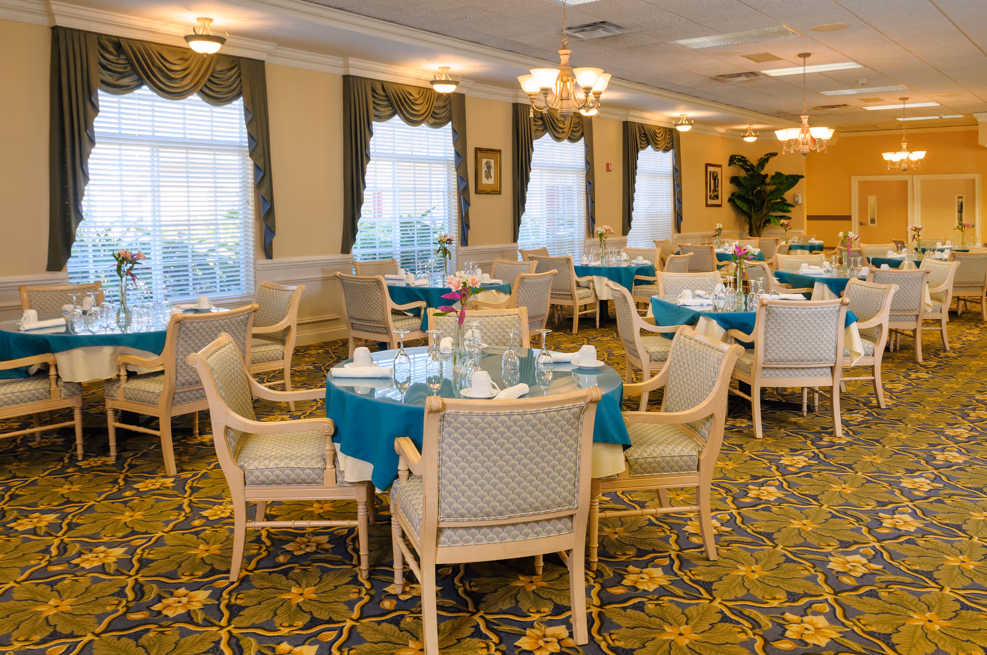 Spacious dining room with multiple round tables set with teal tablecloths, place settings, and upholstered chairs.