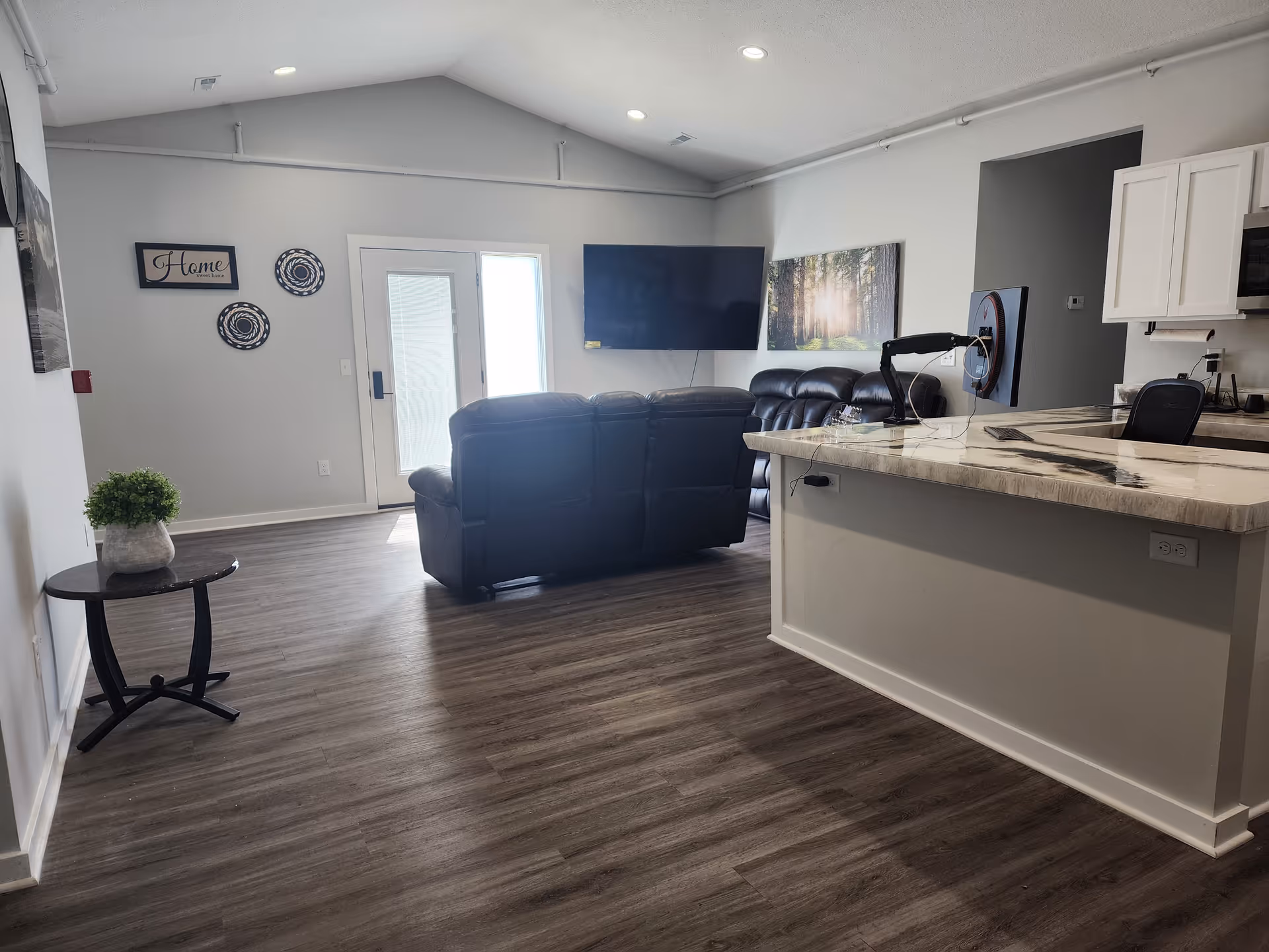 Open-plan living area with leather sofas facing a wall-mounted TV, a kitchen island with marble countertop, and wood-look flooring.