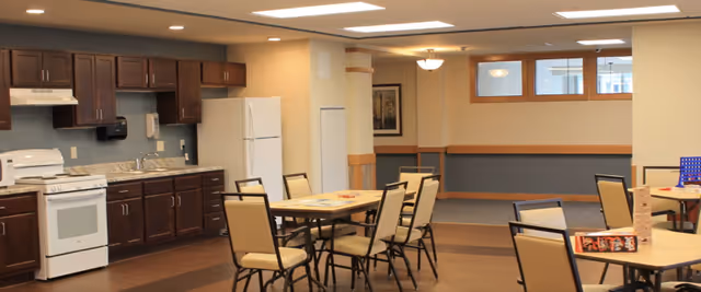 Open communal dining/activity room with tables and chairs and a kitchenette featuring a stove, refrigerator, and dark wood cabinets.
