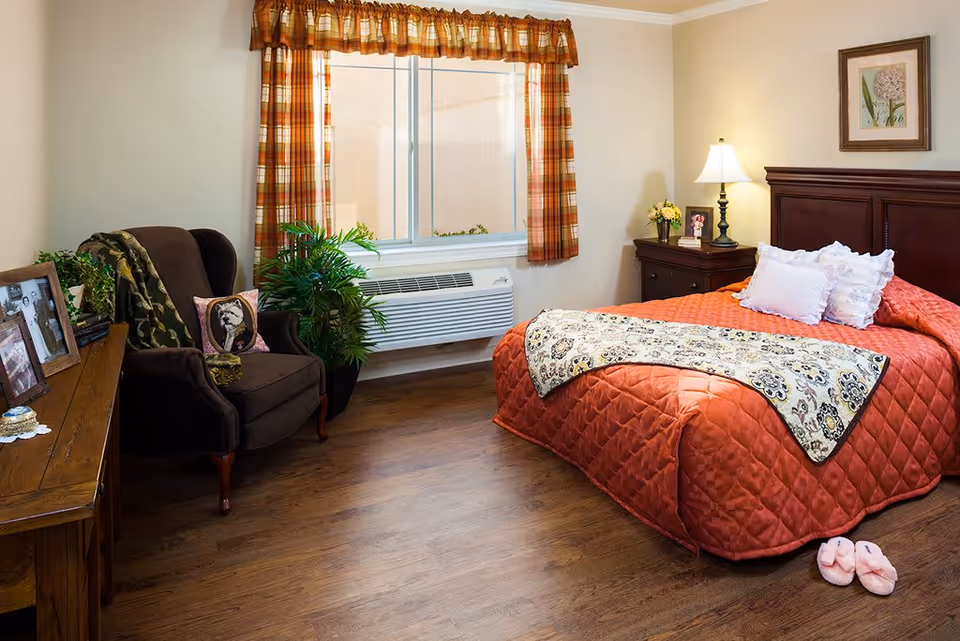 A cozy bedroom with a large bed covered in an orange quilt and decorative pillows. There is a wooden nightstand with a lamp and framed photo, a dark upholstered armchair with a dog-themed pillow, a wooden dresser with framed pictures, a potted plant, and a window with plaid curtains. Pink slippers are placed on the wooden floor near the bed.