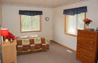 A simple bedroom with two windows covered by dark blue valances, a bed with a patchwork quilt, a wooden nightstand with a red lamp and clock, and a wooden dresser with a vase of red flowers.