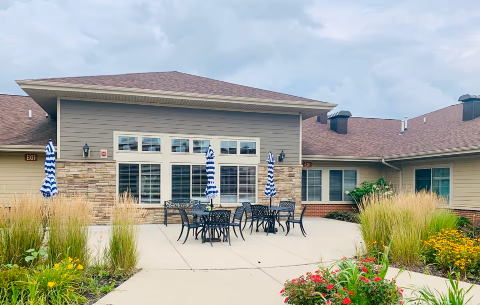 Outdoor patio with metal tables and blue-and-white striped umbrellas surrounded by tall grasses and flowers in front of a single-story building.