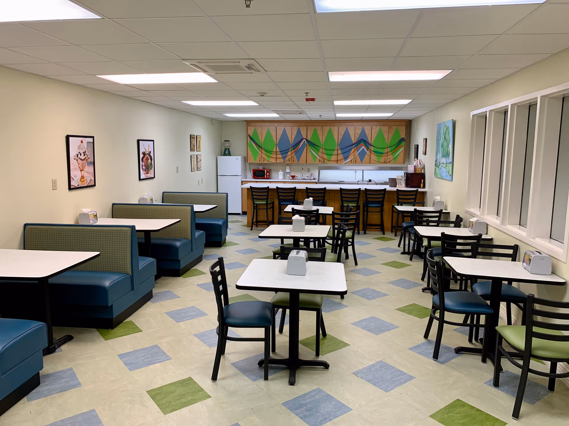 Bright communal dining room with booths, tables and chairs facing a counter and kitchenette with a colorful mural.